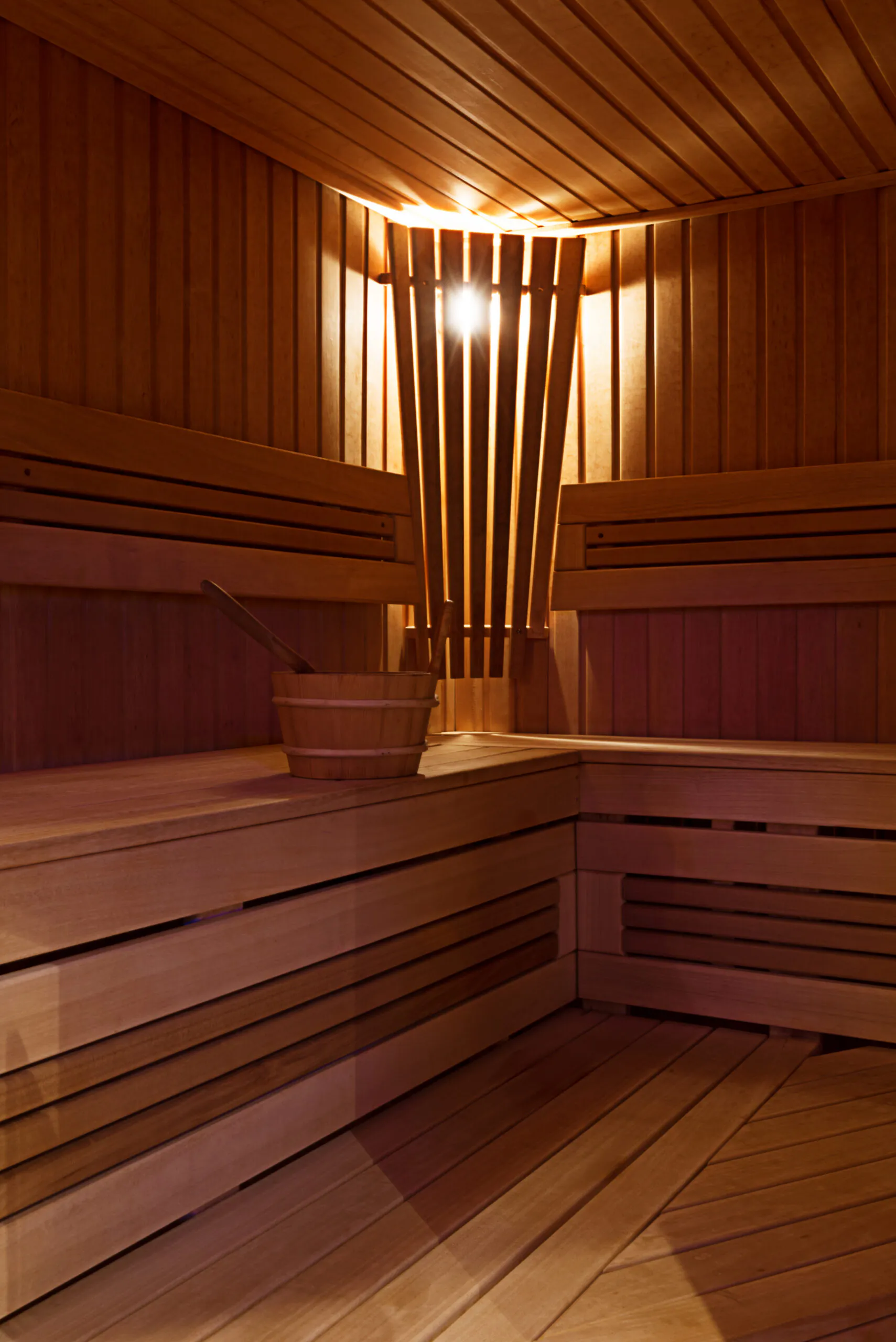 Intérieur d’un sauna en bois clair avec bancs en L, éclairage d’ambiance et seau traditionnel