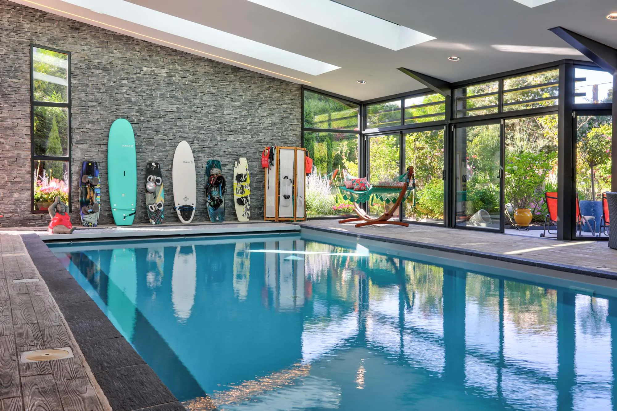 Piscine intérieure rectangulaire avec baies vitrées, mur en pierre et planches de surf